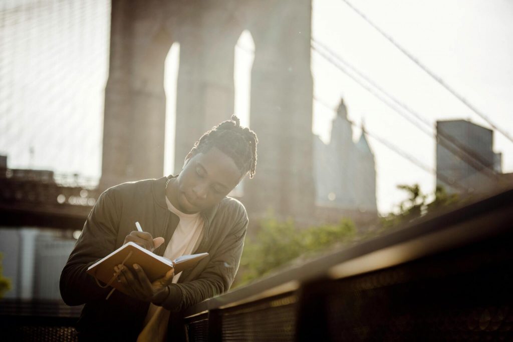 Black thoughtful guy writing in notebook leaning on fence against bridge.
